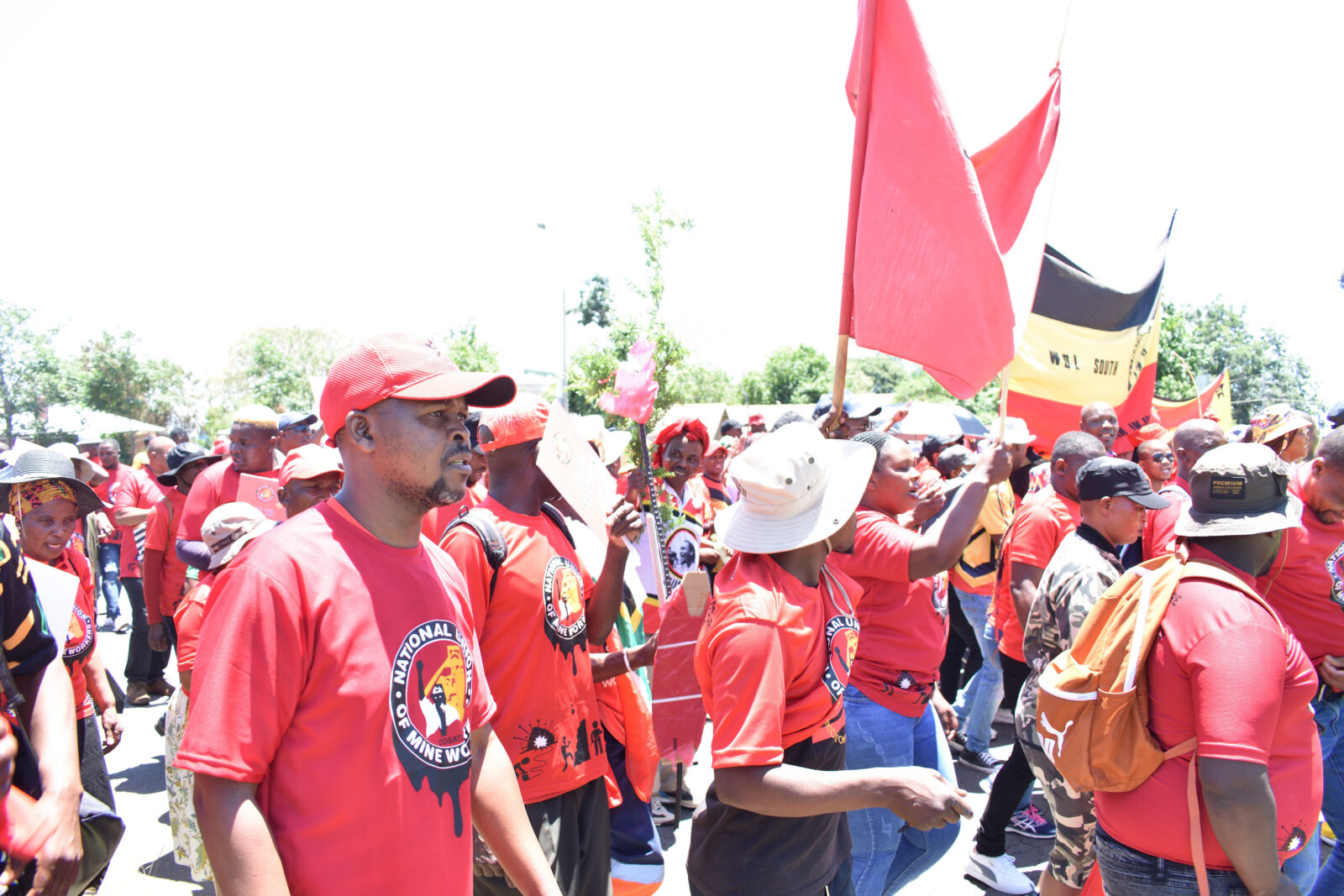 A crowd of NUM (National Union of Mineworkers) members in matching red T-shirts marching through a street in Carletonville during a health and safety march in March 2023, carrying red flags and placards under a bright sunny sky.