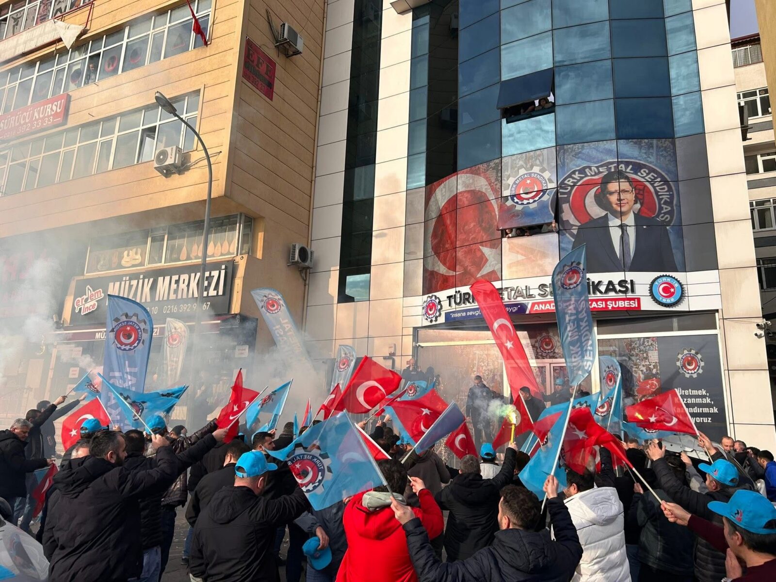 A crowd of workers waving Turkish and Türk Metal Sendikası union flags with smoke flares in the air, gathered in front of the union's Anadolu Branch office building featuring a large portrait of a union leader. 