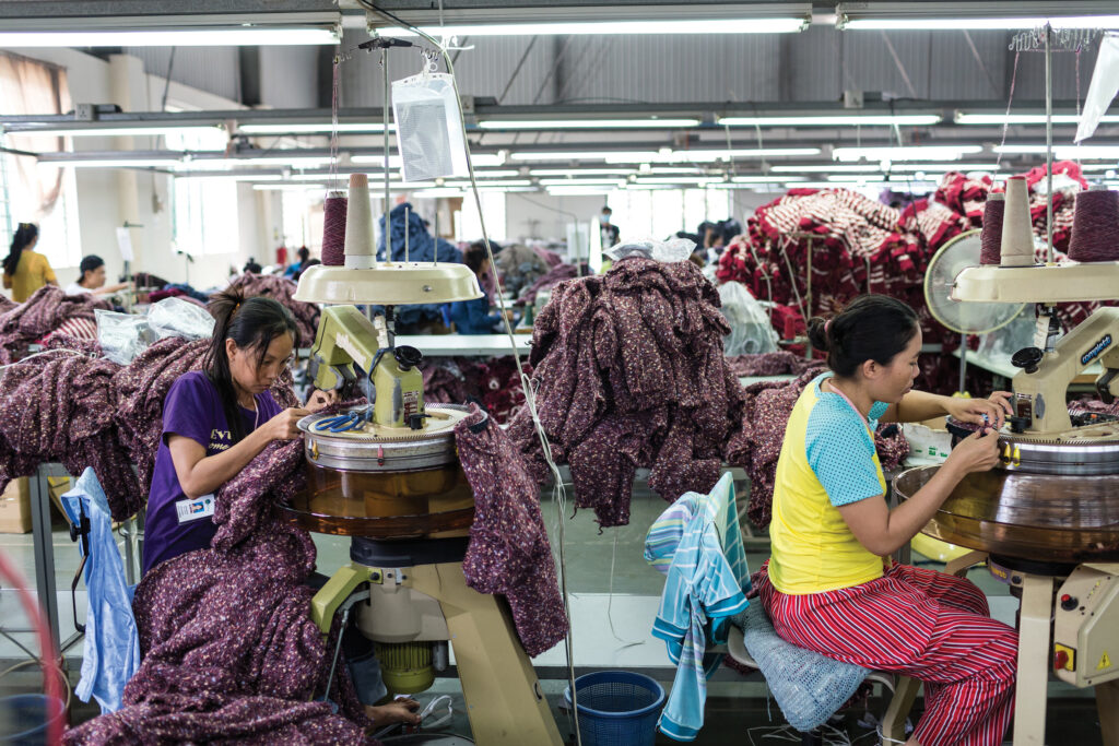 Two women workers at sewing machines on a garment factory floor in Cambodia, surrounded by piles of fabric