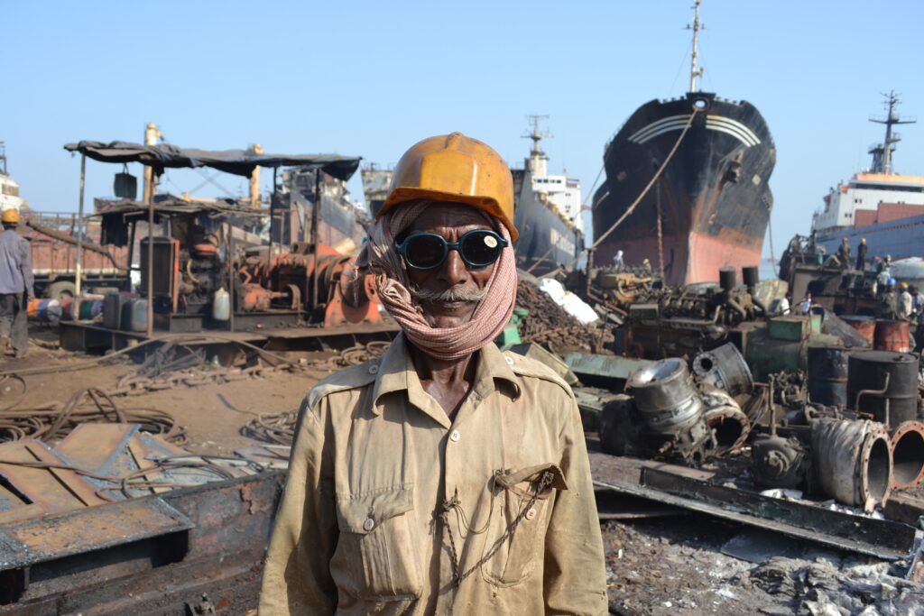 A shipbreaking worker wearing a hard hat and protective goggles stands in front of vessels being dismantled at a shipbreaking yard in Bangladesh