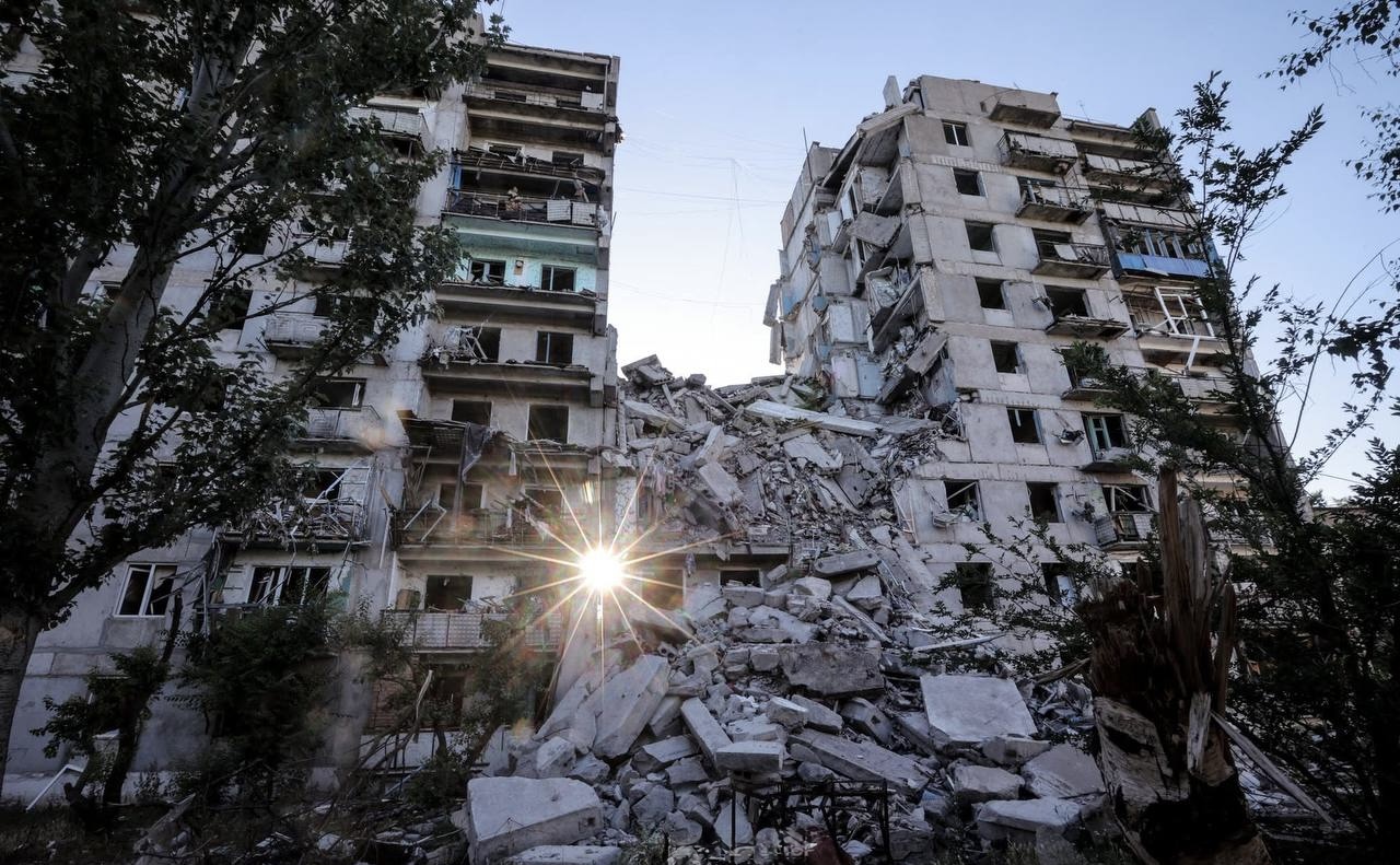 A partially collapsed residential building in Ukraine, with rubble piled at its base and the sun visible through the destruction
