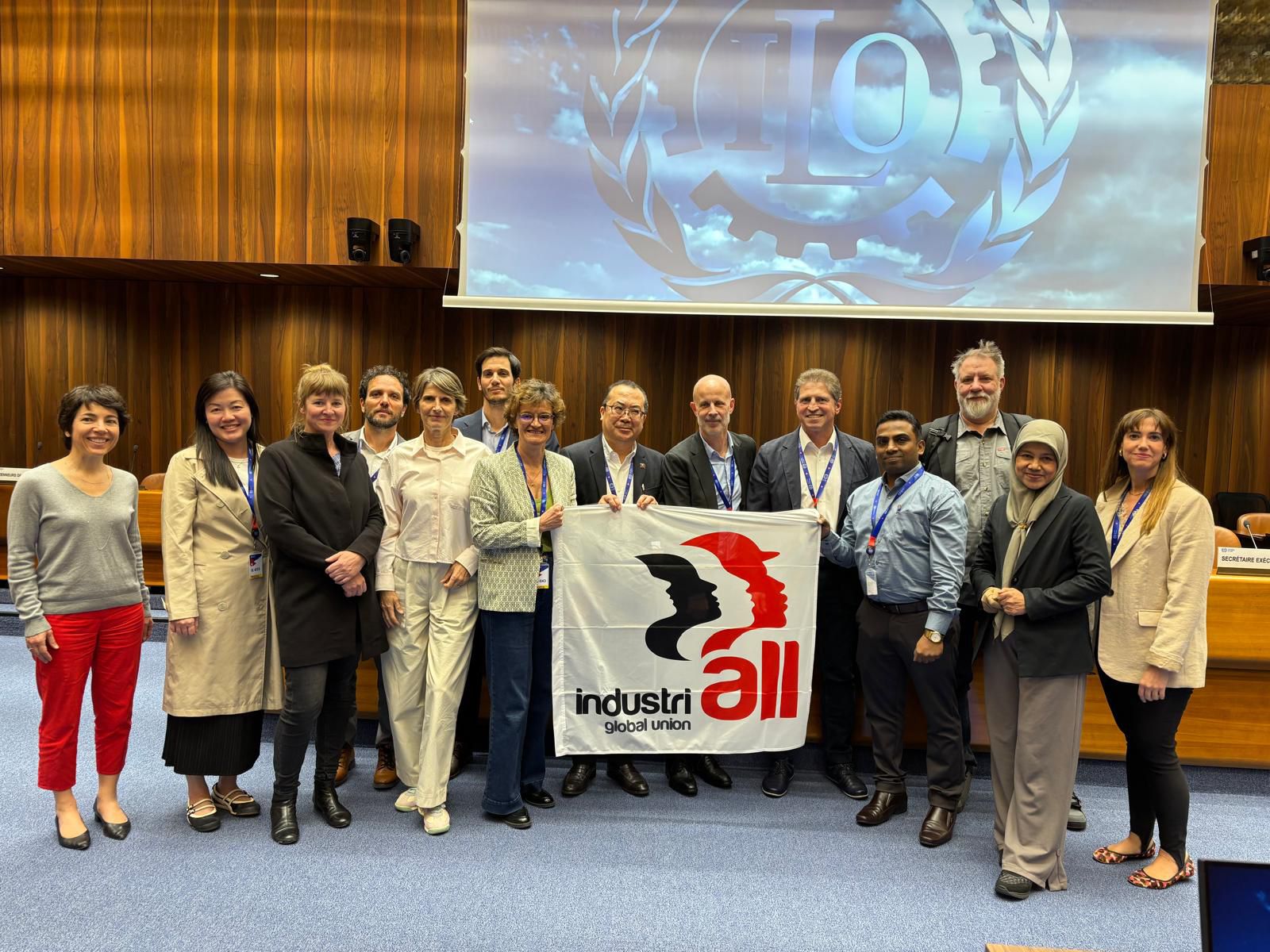 Group photo of IndustriALL Global Union delegates holding the organization's banner inside an ILO conference room in Geneva, with the ILO logo projected on screen in the background, taken during a 2026 event on AI in manufacturing
