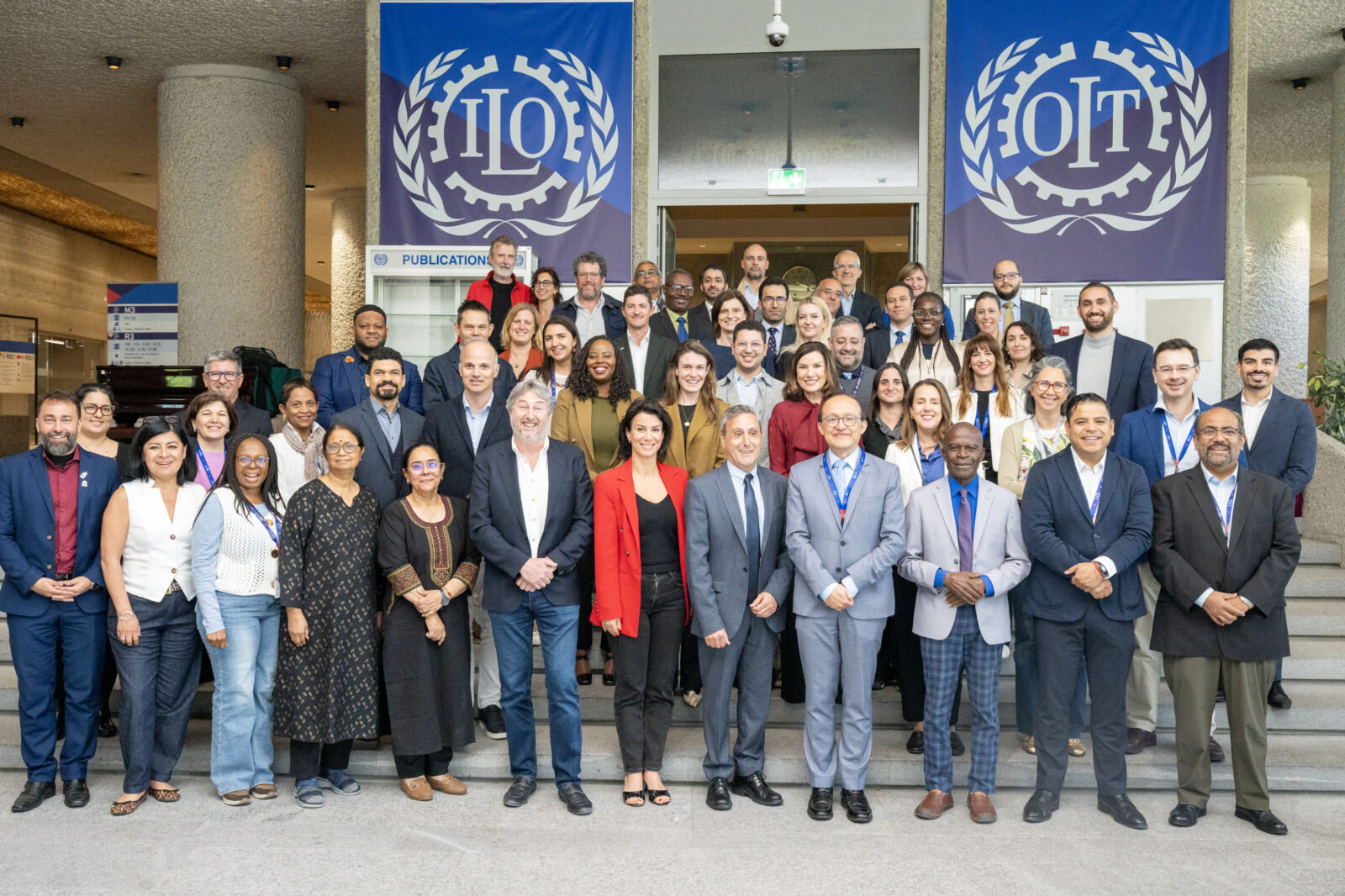 A large group of participants pose for a photo on the steps of the ILO headquarters in Geneva, with the ILO and OIT logos visible on banners in the background.