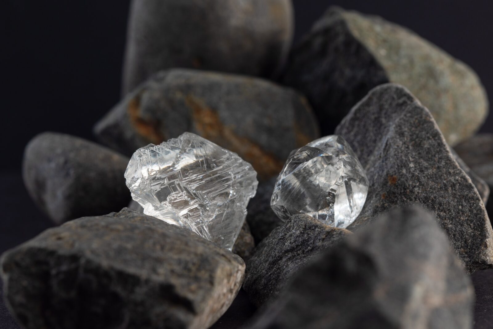 Two uncut natural diamonds resting among grey rough rocks against a dark background