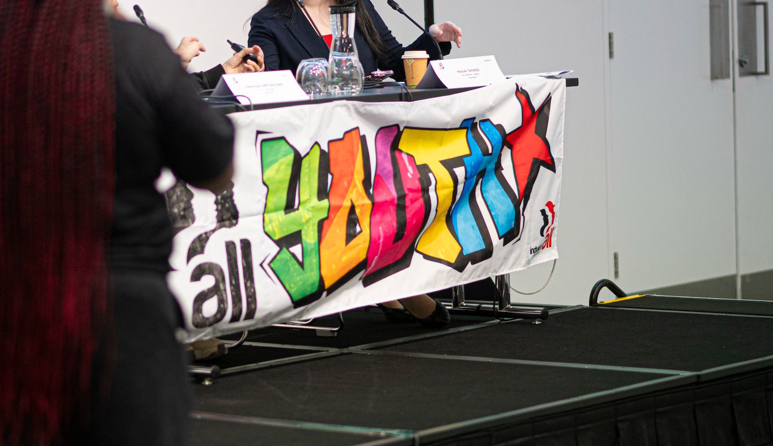 A conference panel table on a raised stage displaying a colourful graffiti-style "all youth" banner with the IndustriALL logo, with two participants seated behind it.