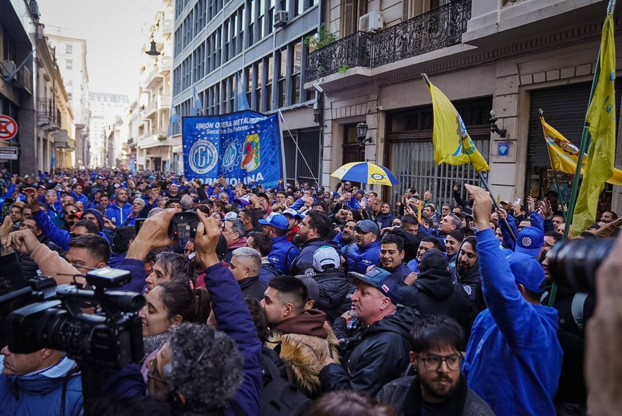 A large crowd of metalworkers fills a city street in Argentina, many wearing blue union jackets and caps, with UOM (Unión Obrera Metalúrgica) banners and yellow and blue flags visible above the crowd.