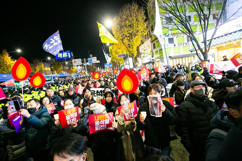 A large crowd of workers at a night-time protest in Seoul, holding red placards and flame-shaped signs with union flags visible in the background.