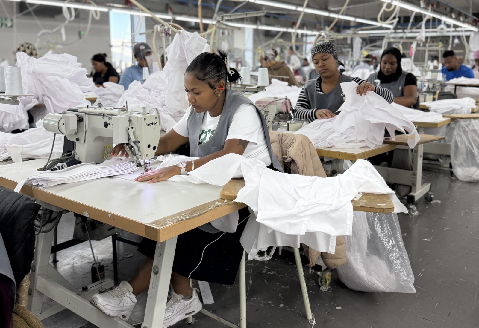 Women garment workers sewing and inspecting white fabric at industrial sewing machines on a factory floor at a Pep Clothing facility in Cape Town, South Africa, April 2026.