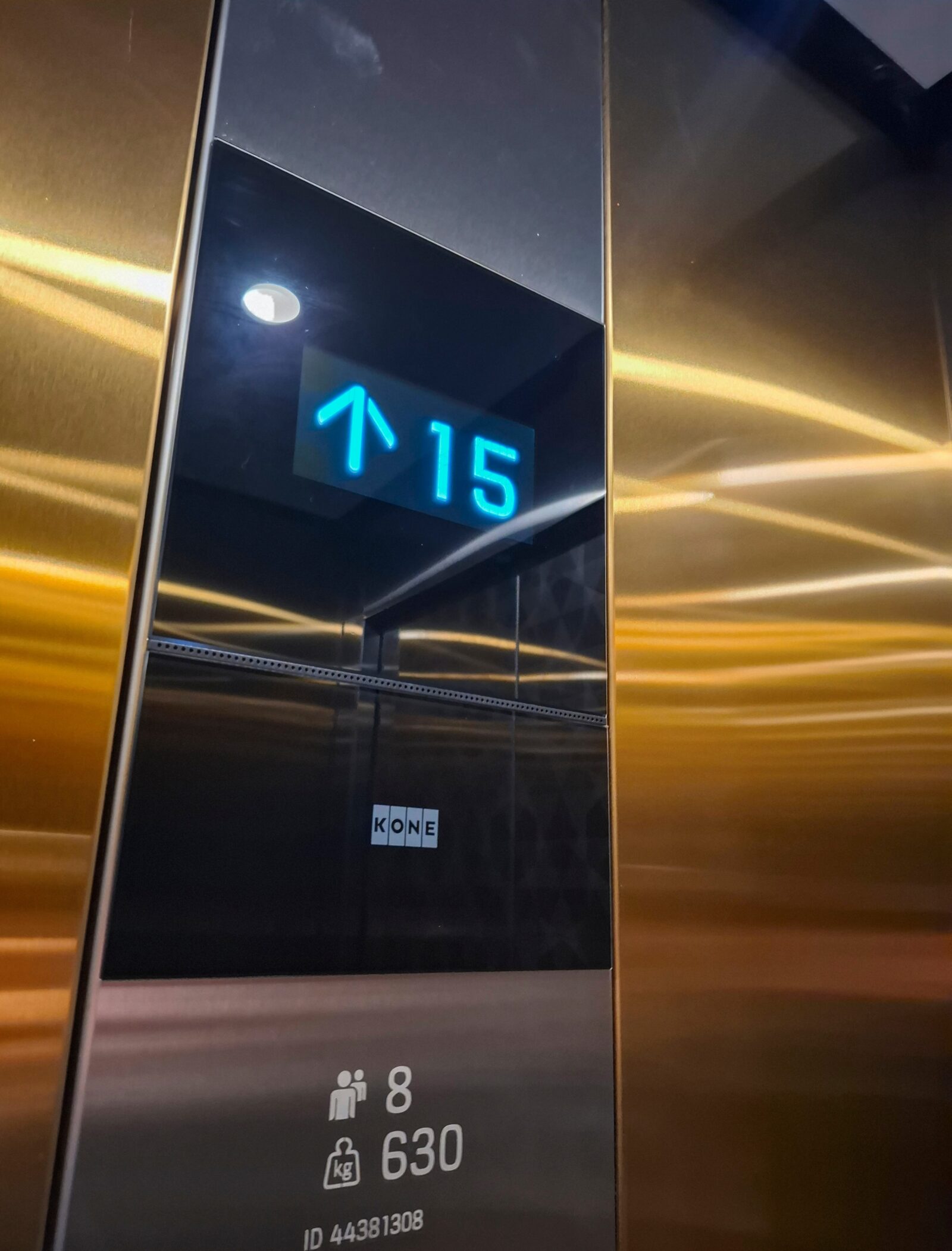 Interior panel of a KONE elevator showing a cyan digital display with an upward arrow and the number 15, with the KONE logo and capacity information below, set against gold-toned brushed metal walls.