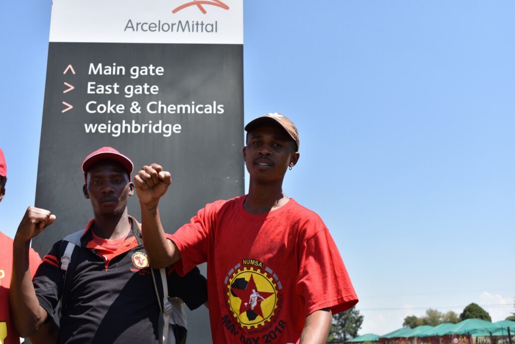 Two workers wearing red and black union shirts raise their fists in front of an ArcelorMittal facility entrance sign in South Africa. 