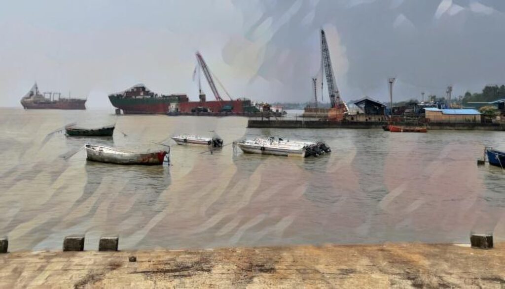 Small boats moored in the foreground of a shipbreaking yard in Bangladesh, with large vessels and industrial cranes visible in the background under a hazy sky