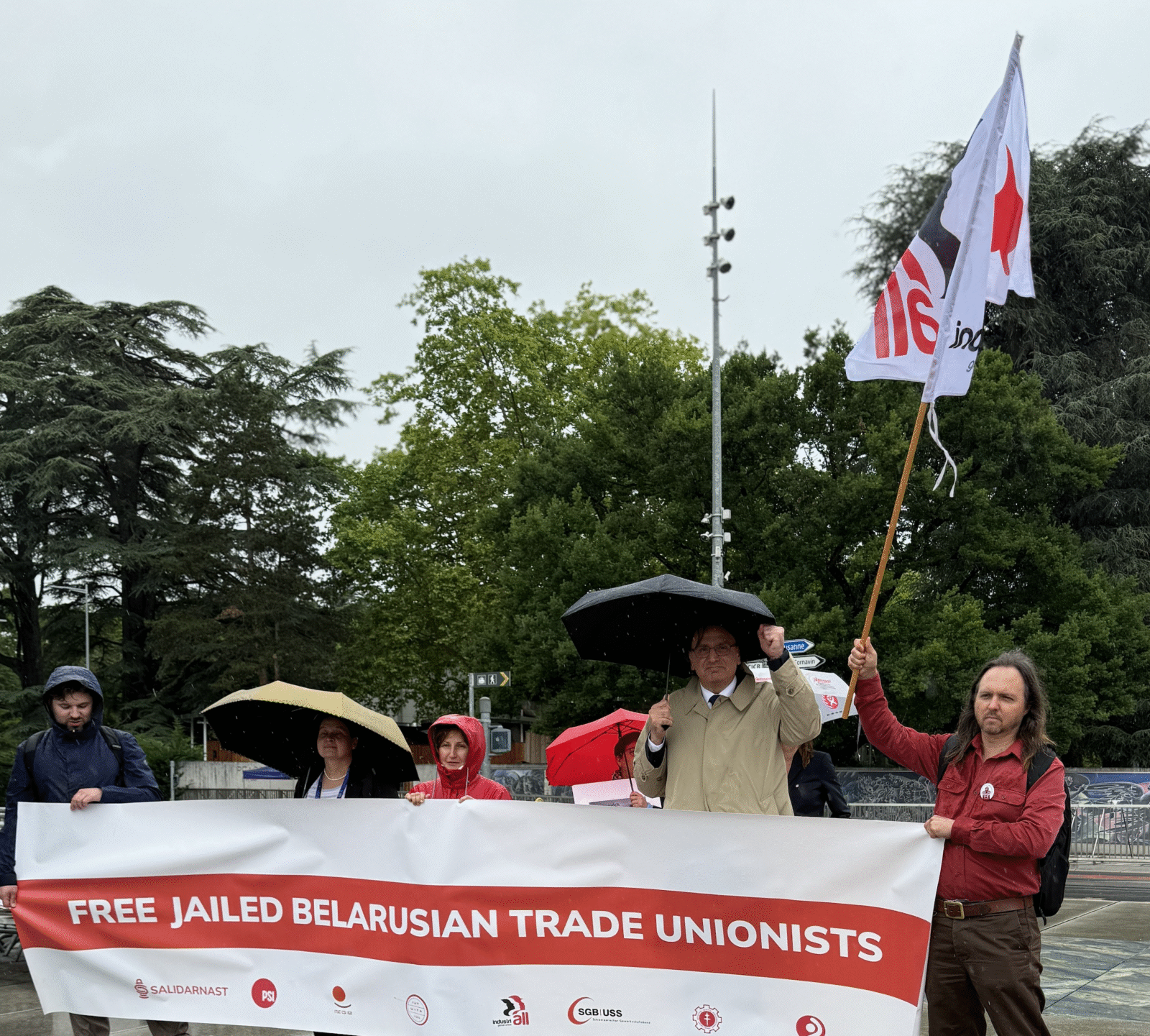 Sindicalistas sosteniendo una pancarta «Free Jailed Belarusian Trade Unionists» frente al monumento de la Silla Rota en Ginebra, con una bandera de IndustriALL levantada en alto, durante una manifestación bajo la lluvia
