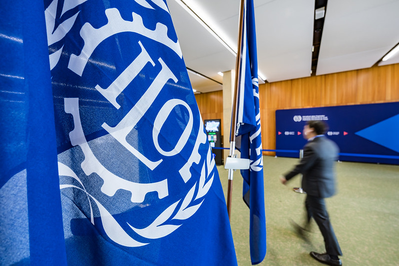 ILO flags in the foreground with a delegate walking past a blue ILO backdrop at the 114th International Labour Conference, Geneva