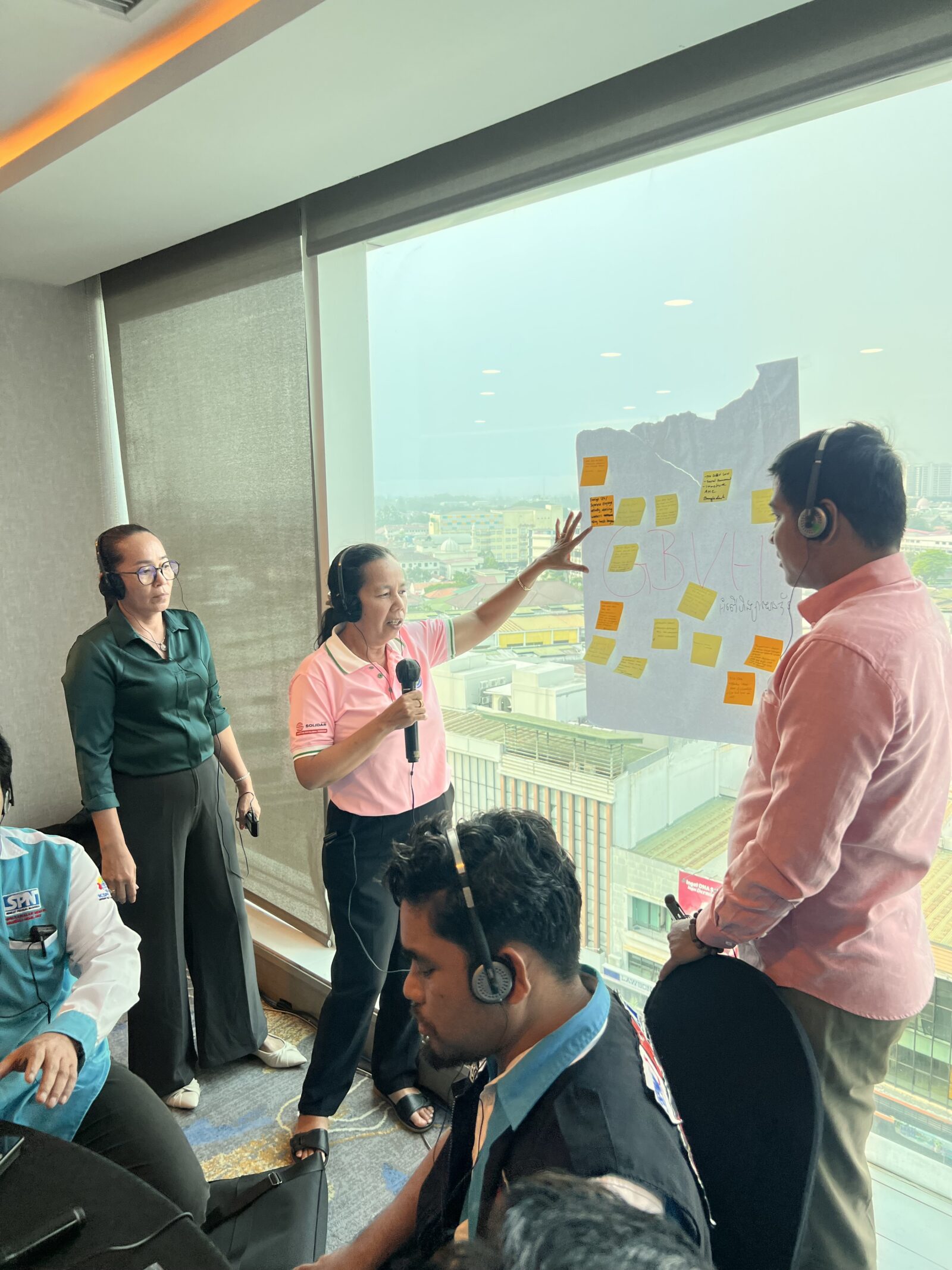 Woman in pink polo shirt speaks into a microphone and gestures toward a flip chart labeled "GBVH" covered in sticky notes, while other workshop participants listen wearing headphones.