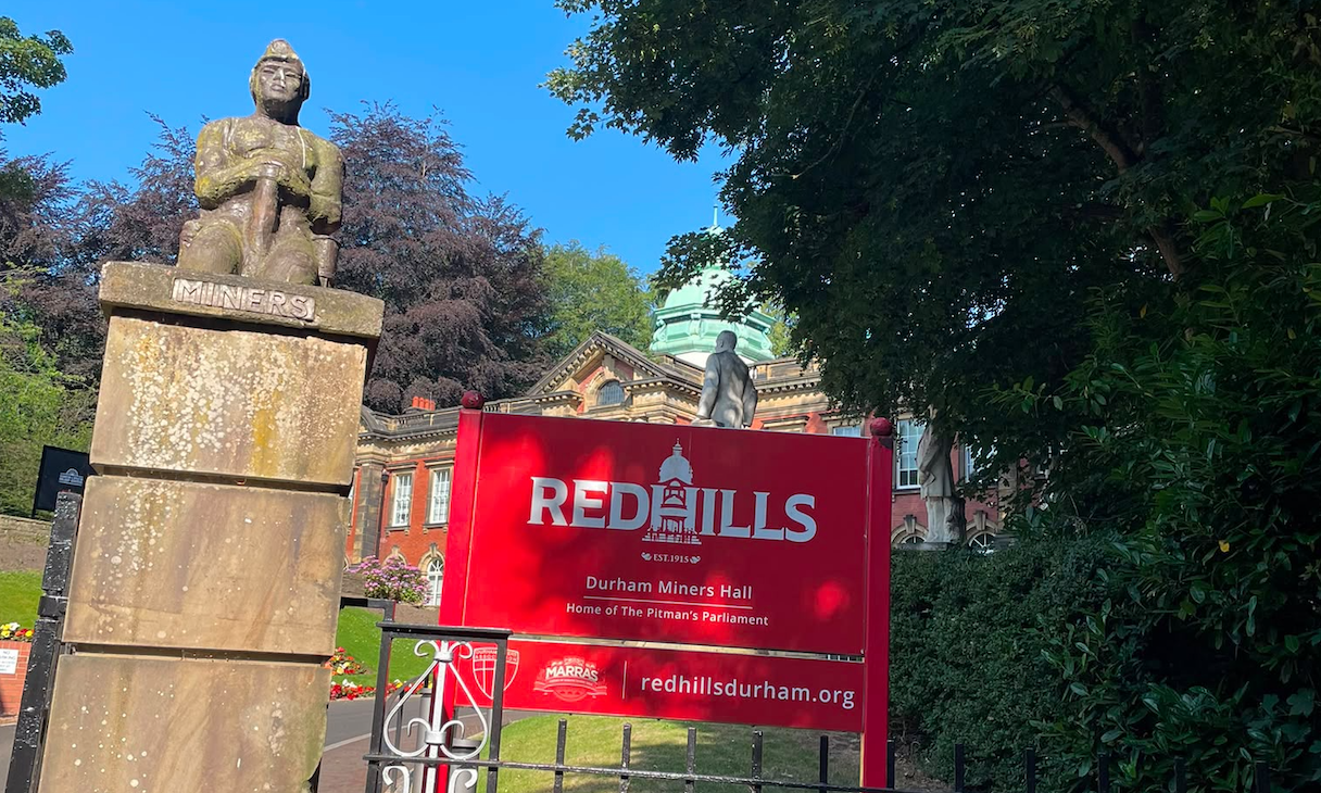 A weathered stone statue of a miner atop a plinth reading "Miners", beside a red Redhills sign in the grounds of Durham Miners' Hall, with the hall's green dome visible behind.