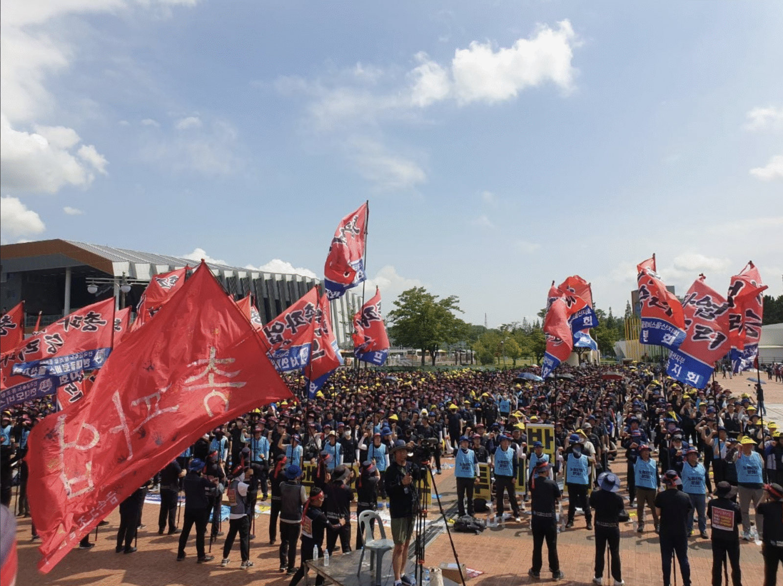 Thousands of Korean Metal Workers' Union members rally outdoors, waving red flags reading 민주노총, with workers in black shirts, hard hats and blue vests filling a large plaza.