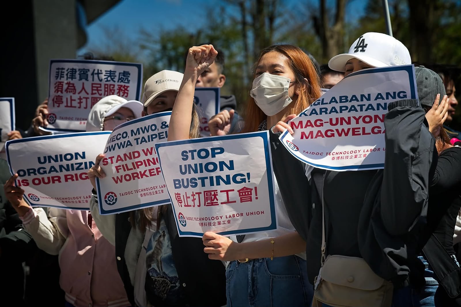 A protester raises her fist while holding a "stop union busting!" sign at a Taidoc Technology Labor Union rally, surrounded by supporters with placards.