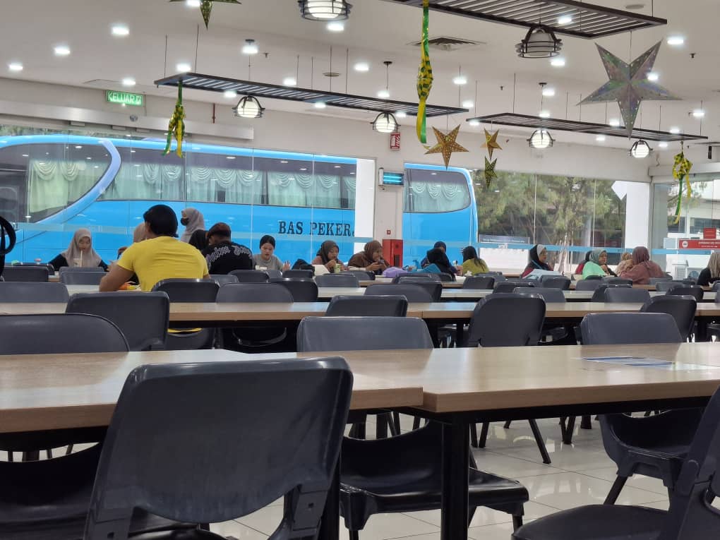 Workers seated at tables in a factory canteen with a company worker bus visible through the windows outside.