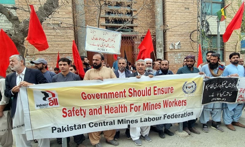 A crowd of men holding red flags and a white banner reading "Government Should Ensure Safety and Health For Mines Workers, Pakistan Central Mines Labour Federation," outside a building with an Urdu-language protest sign.
