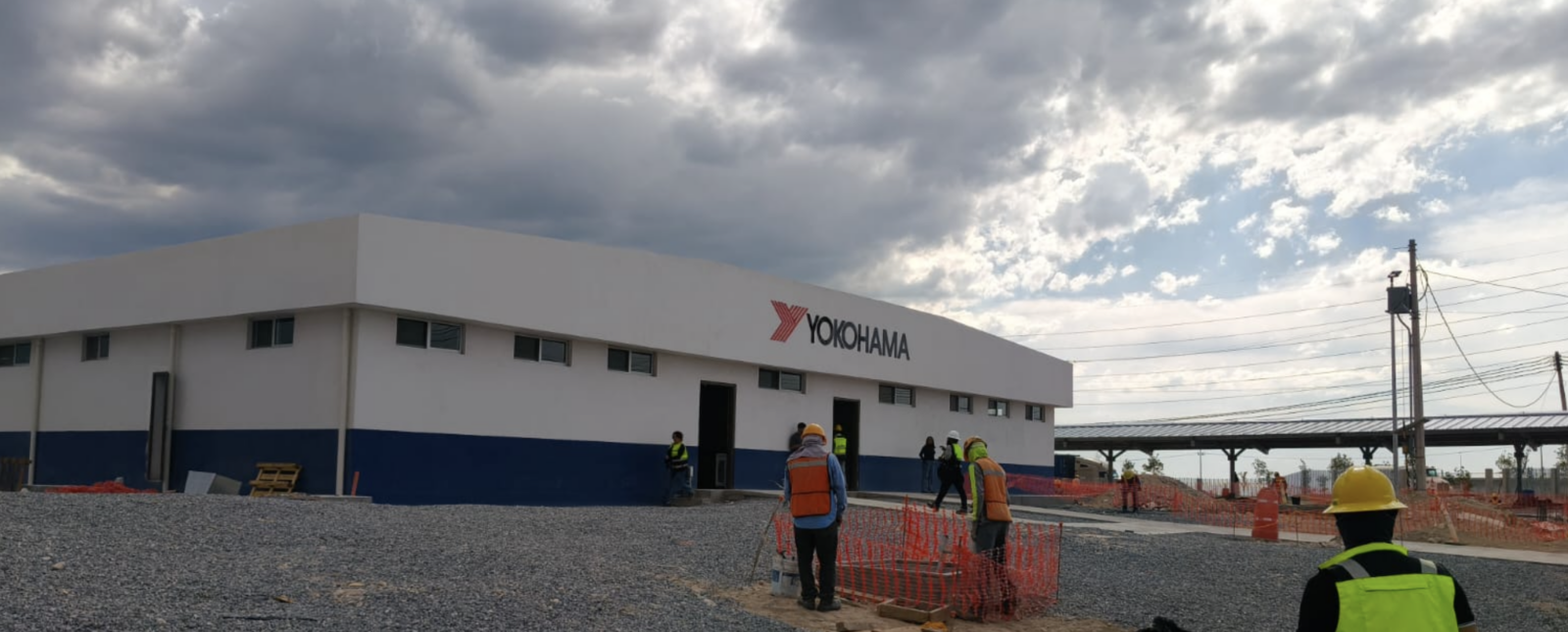 Workers in hi-vis vests and hard hats outside the newly built Yokohama tyre plant in Mexico, with solar panel canopies visible in the background under a cloudy sky.