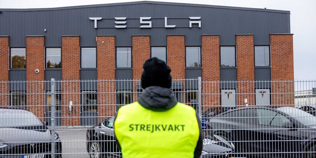 A picket guard in a yellow "strejkvakt" vest stands outside a fenced Tesla building in Sweden.