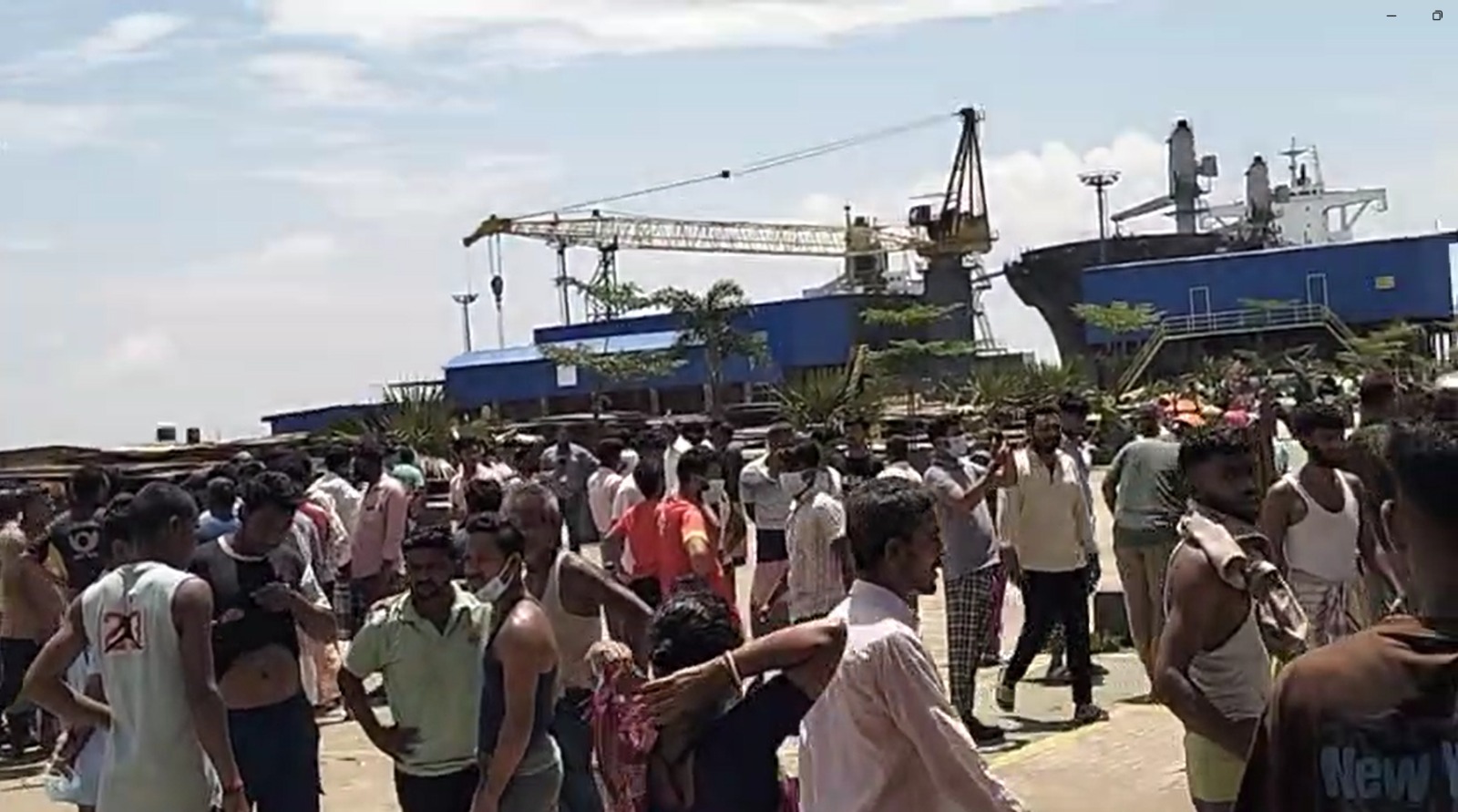 a crowd of shipyard workers stands in an open yard in Bangladesh, with a partially dismantled ship and dockside crane visible behind them, on the day of the fatal gas exposure incident.