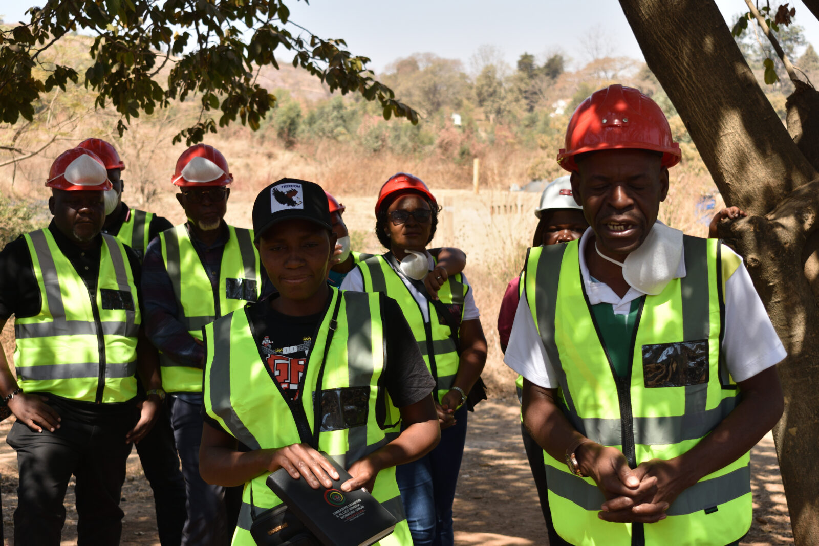 Mineworkers wearing red hard hats, high-visibility vests and dust masks stand together outdoors near a mine site, with one holding a folder branded "Zimbabwe Diamond and Allied Minerals Workers Union".