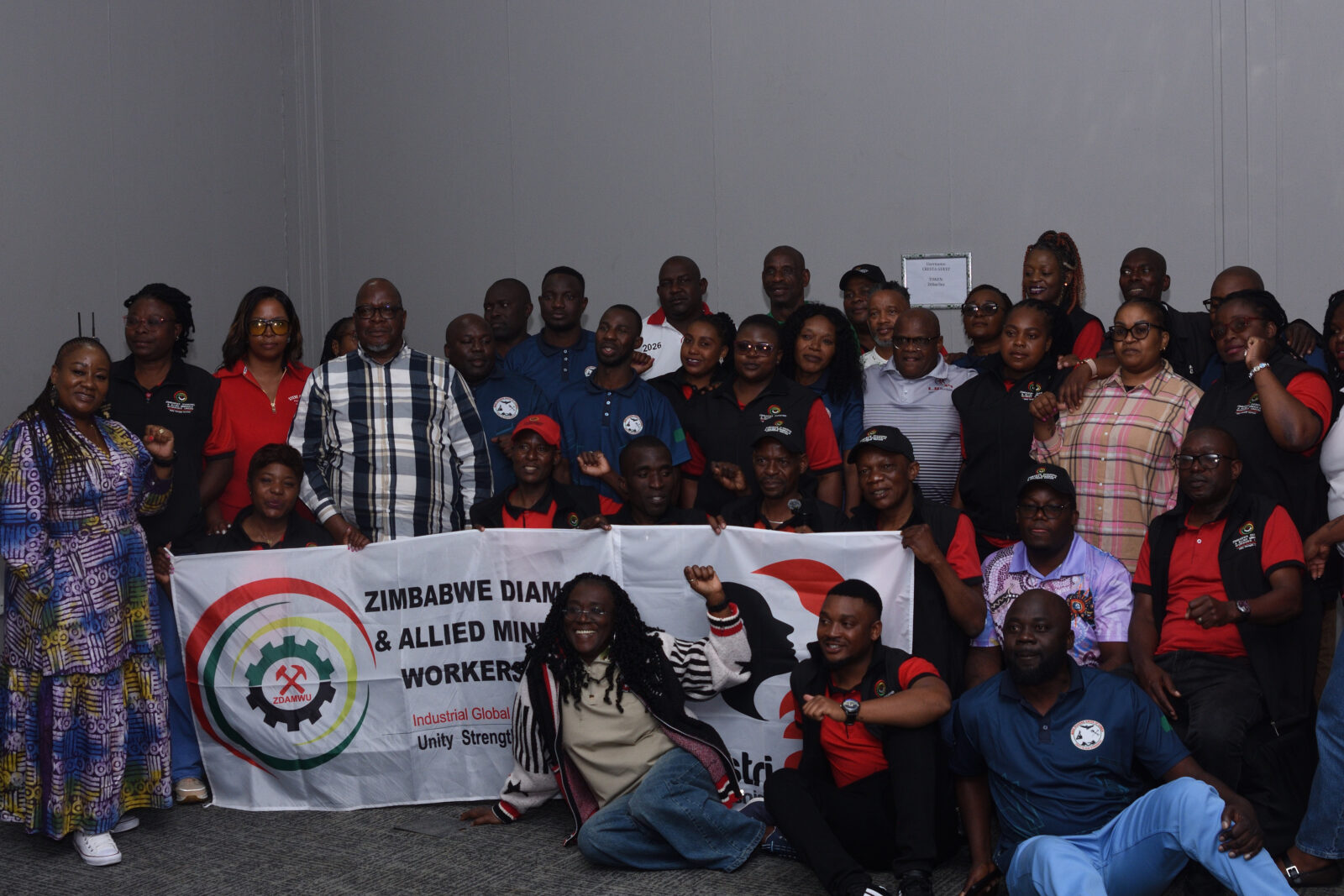 A group photo of around 40 people, many in ZDAMWU-branded red and black shirts, standing and kneeling behind a white banner that reads "Zimbabwe Diamond & Allied Minerals Workers Union, Industrial Global, Unity Strength", several with fists raised