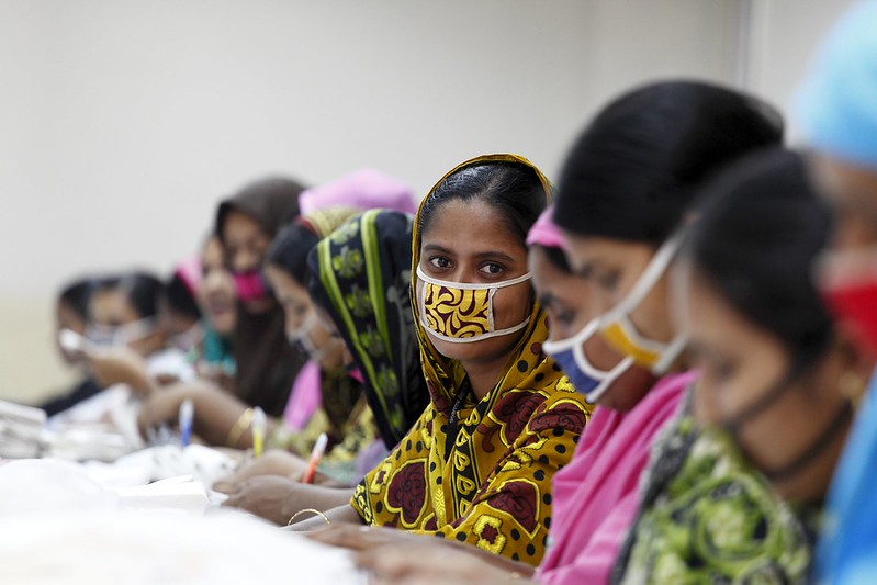 Trabajadoras del sector de la confección sentadas en una mesa durante una sesión de formación en Bangladesh. La mujer del centro mira directamente a la cámara, con una mascarilla de tela estampada y un pañuelo amarillo.