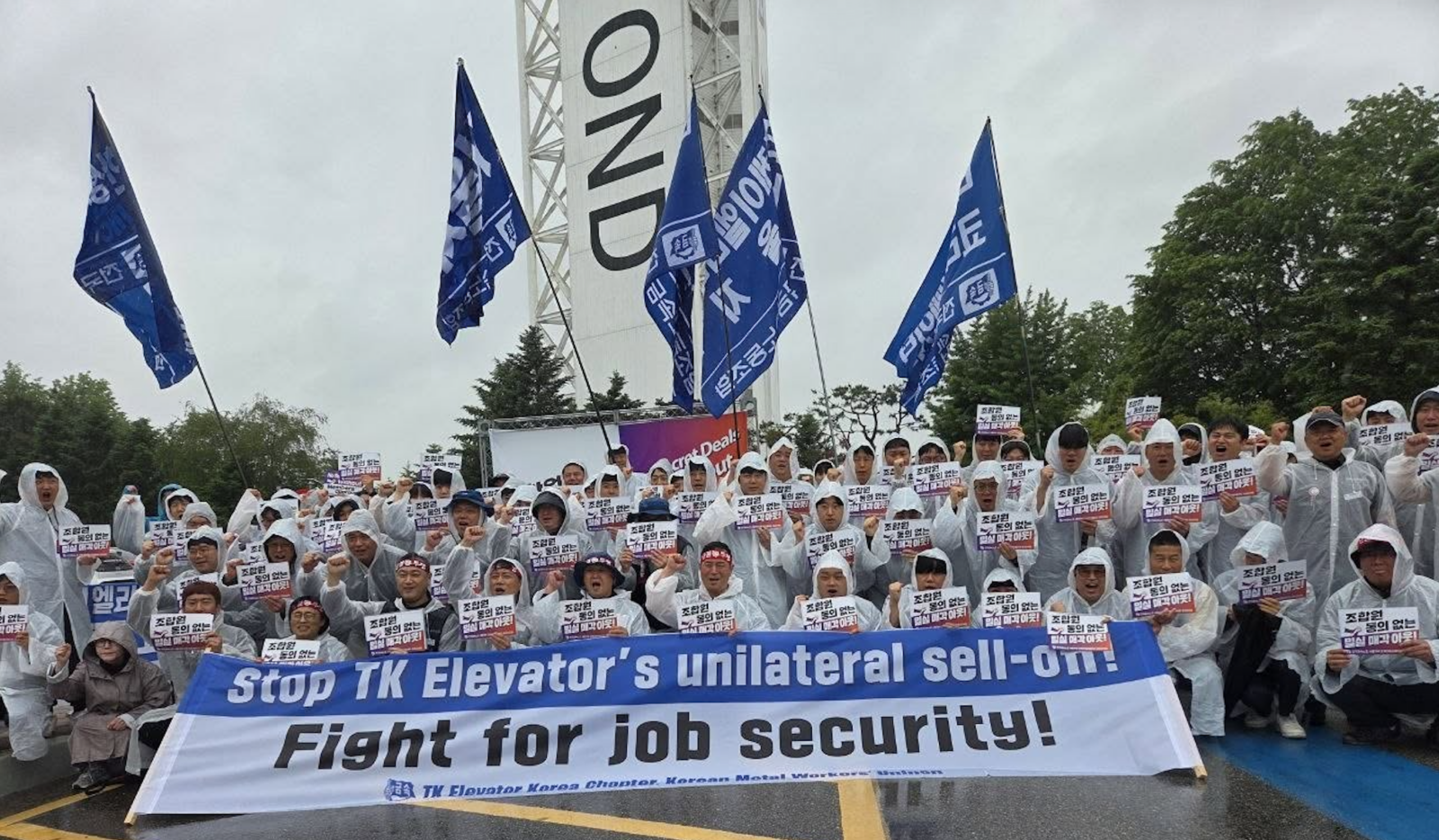Hundreds of workers in white raincoats and caps hold protest signs and raise blue union flags at a rally. A large banner reads: "stop TK Elevator's unilateral sell-on. Fight for job security!"