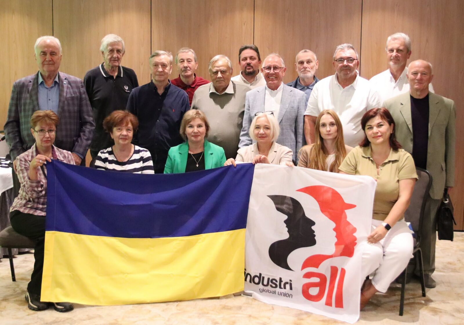 Group photo of Ukrainian trade union delegates standing and seated indoors, with two women in the front row holding up the Ukrainian flag and an IndustriALL Global Union flag.