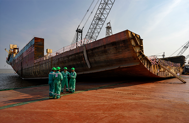 Four workers in protective green uniforms stand on the red earth of a ship recycling yard, dwarfed by a massive vessel undergoing dismantling with industrial cranes and equipment visible above.