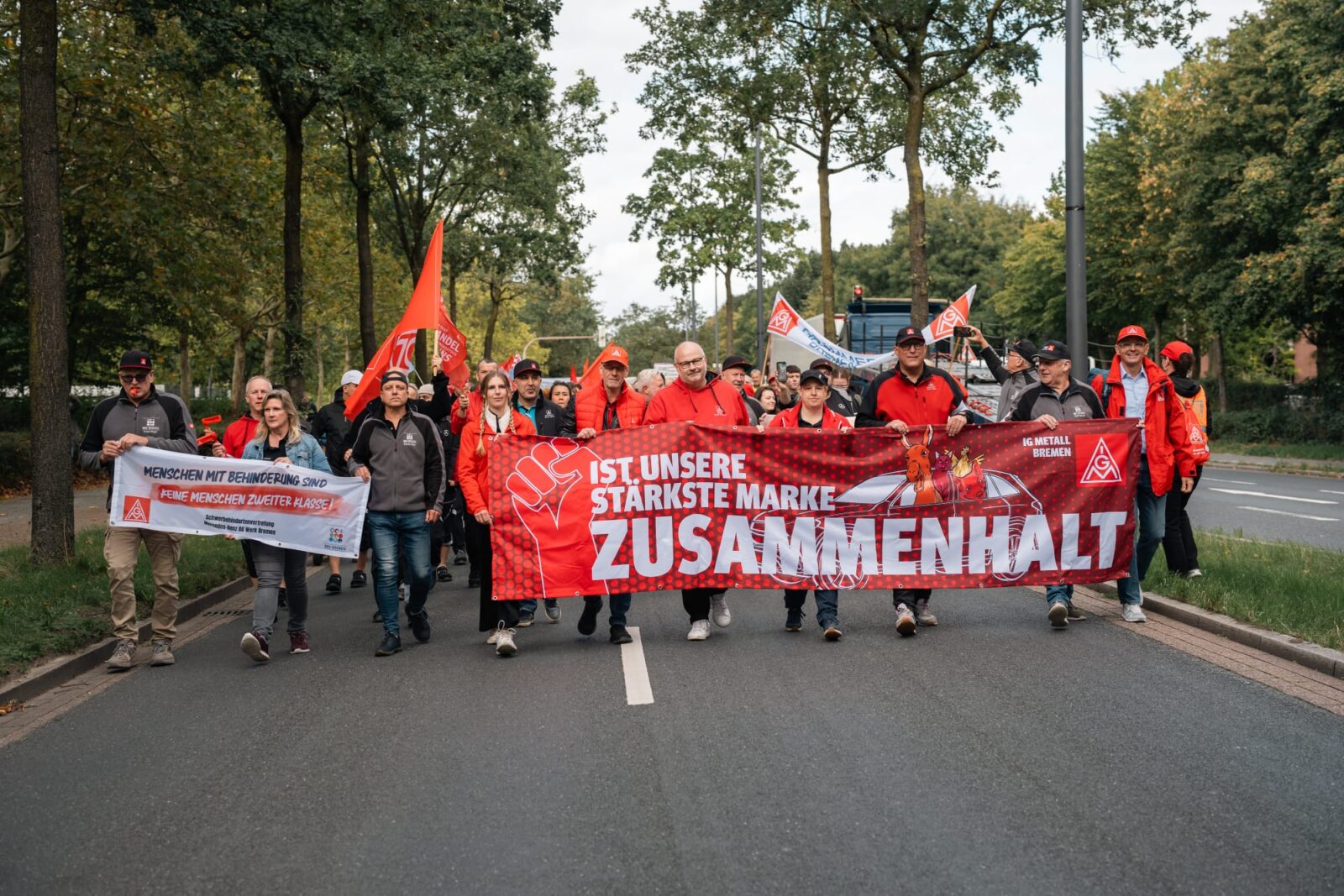 IG Metall Bremen members and Mercedes-Benz workers march down a street holding a red banner reading "Zusammenhalt ist unsere stärkste Marke" (Solidarity is our strongest brand).
