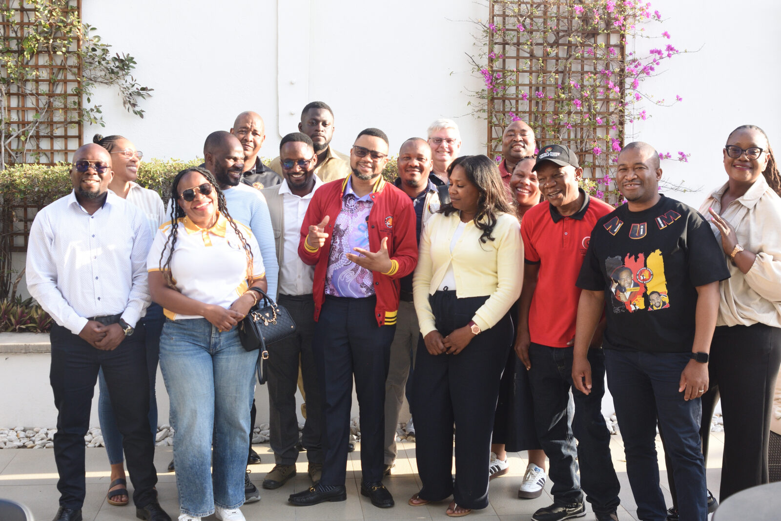 Fourteen trade unionists from Botswana, Lesotho, Namibia, South Africa, Tanzania and Zimbabwe standing together outdoors in a courtyard with purple flowering plants and lattice work behind them