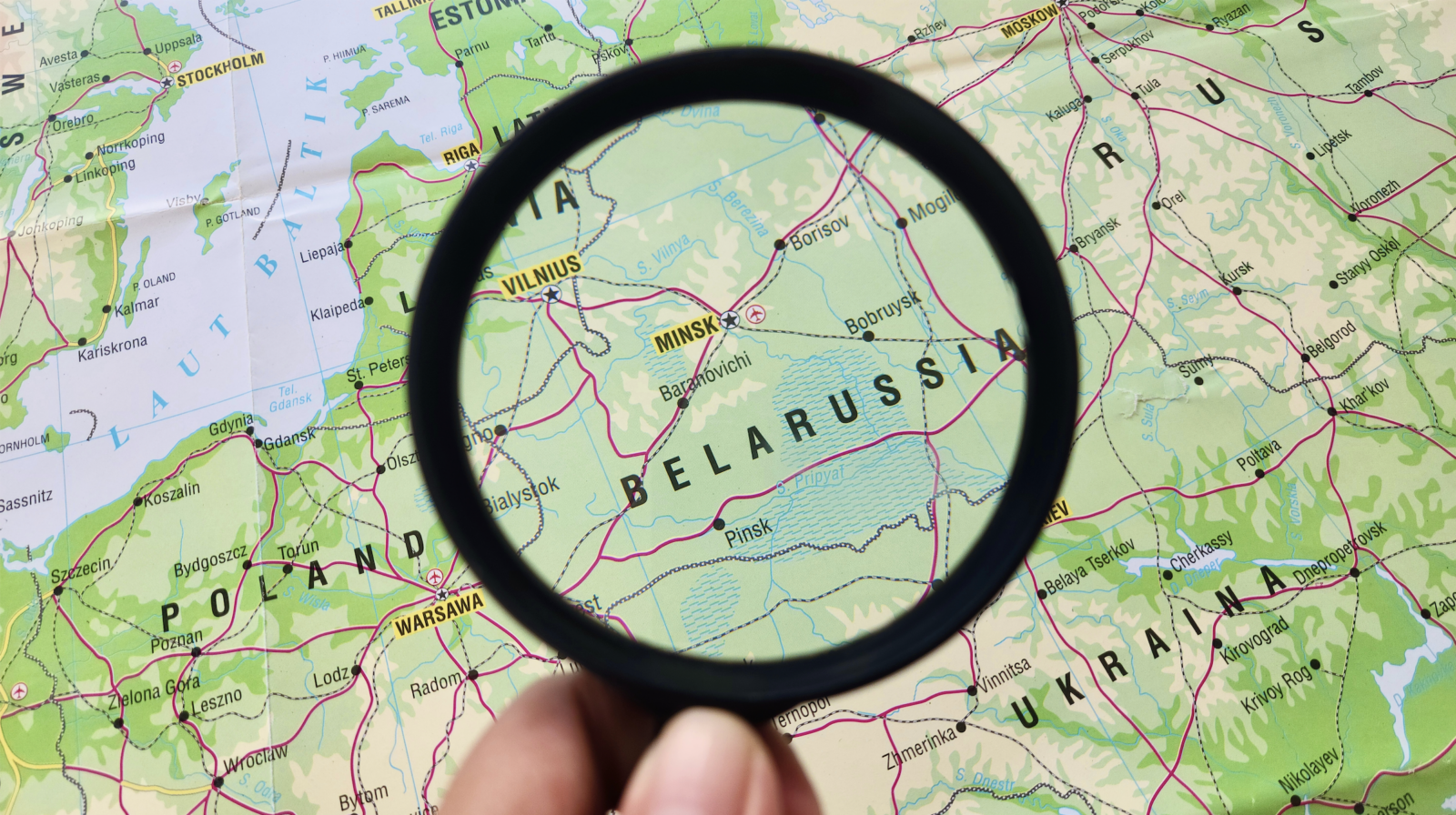 Magnifying glass focusing on Belarus on a physical map of Eastern Europe, with surrounding countries Poland, Lithuania, Latvia and Russia visible.
