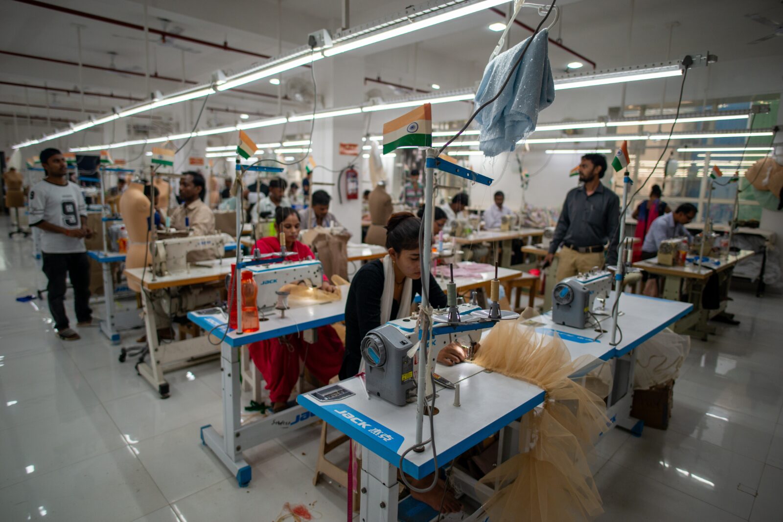 Women sew garments at industrial sewing machines in a large, brightly lit factory in India, while men stand watching nearby. Small Indian flags are attached to the machines.
