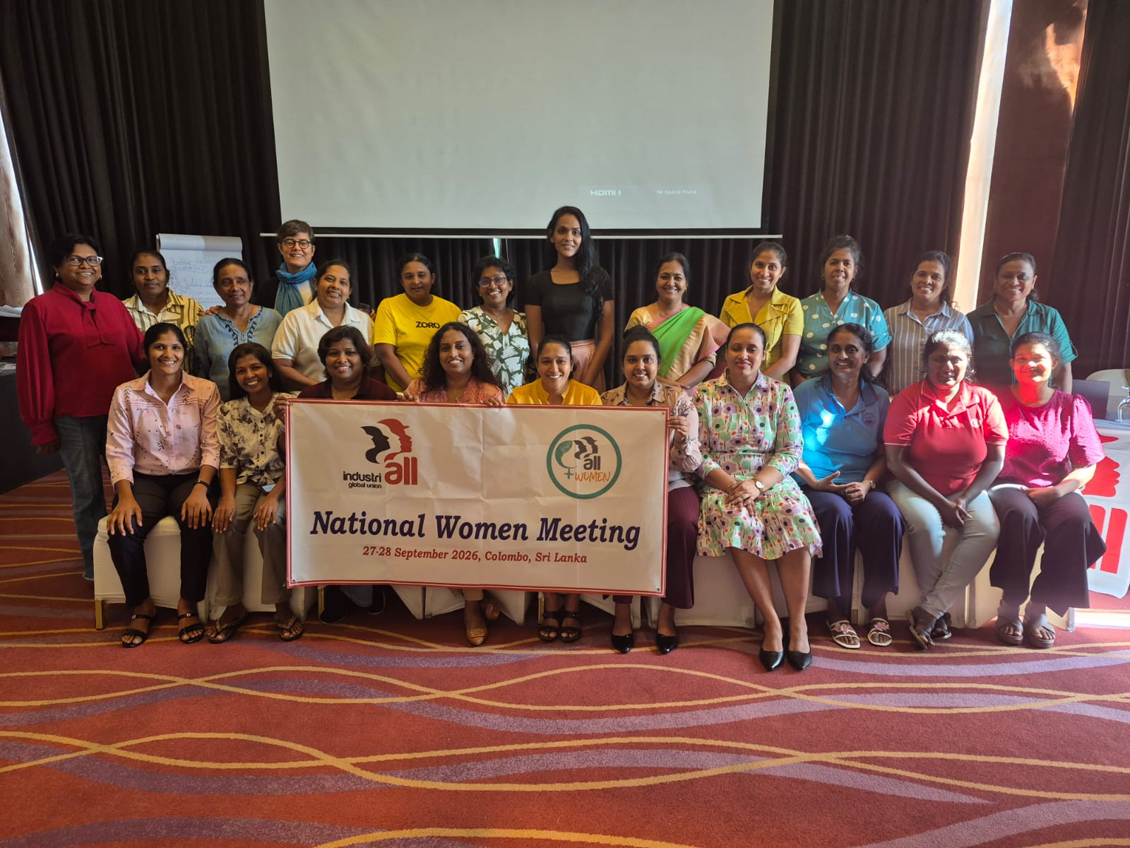 More than 20 women pose for a group photo in a meeting room. Ten sit in a front row holding a white banner, and about 13 stand behind them. The banner shows the IndustriALL Global Union and IndustriALL Women logos and reads “national women meeting, 27-28 September 2026, Colombo, Sri Lanka”