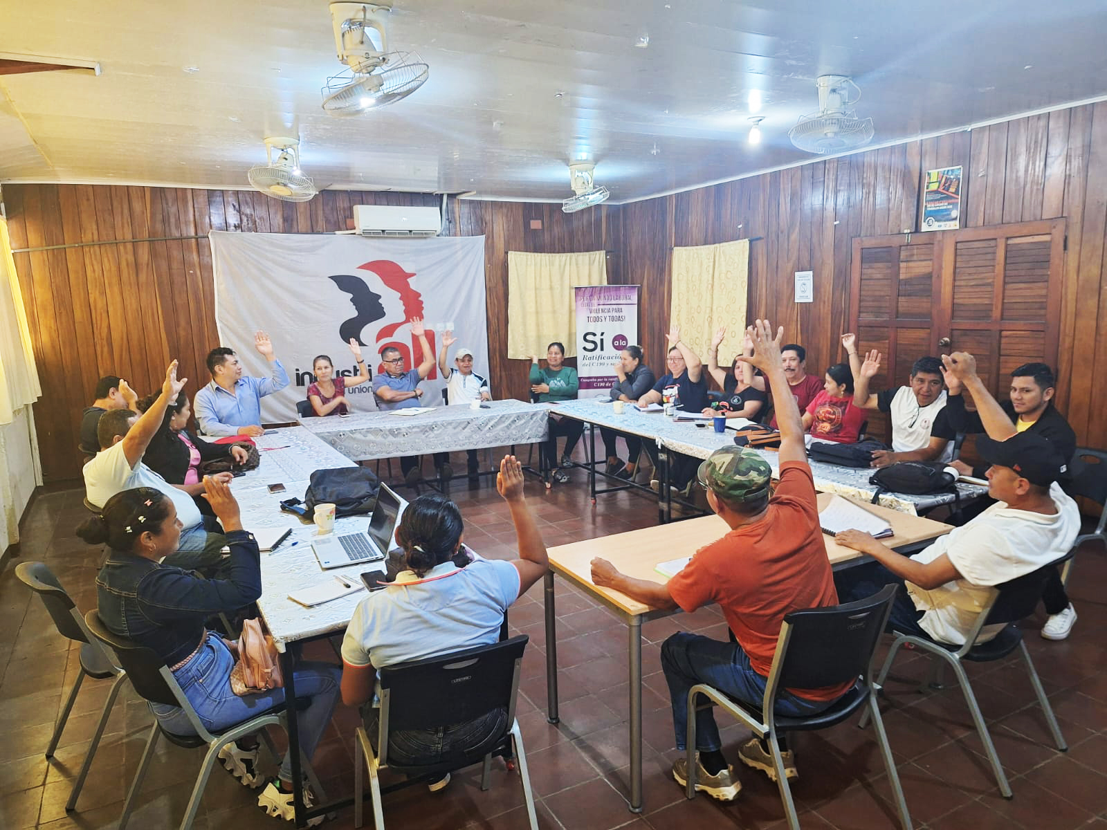 Grupo de personas reunidas alrededor de mesas en una sala de reuniones, participando en una votación a mano alzada frente a un banner sindical.