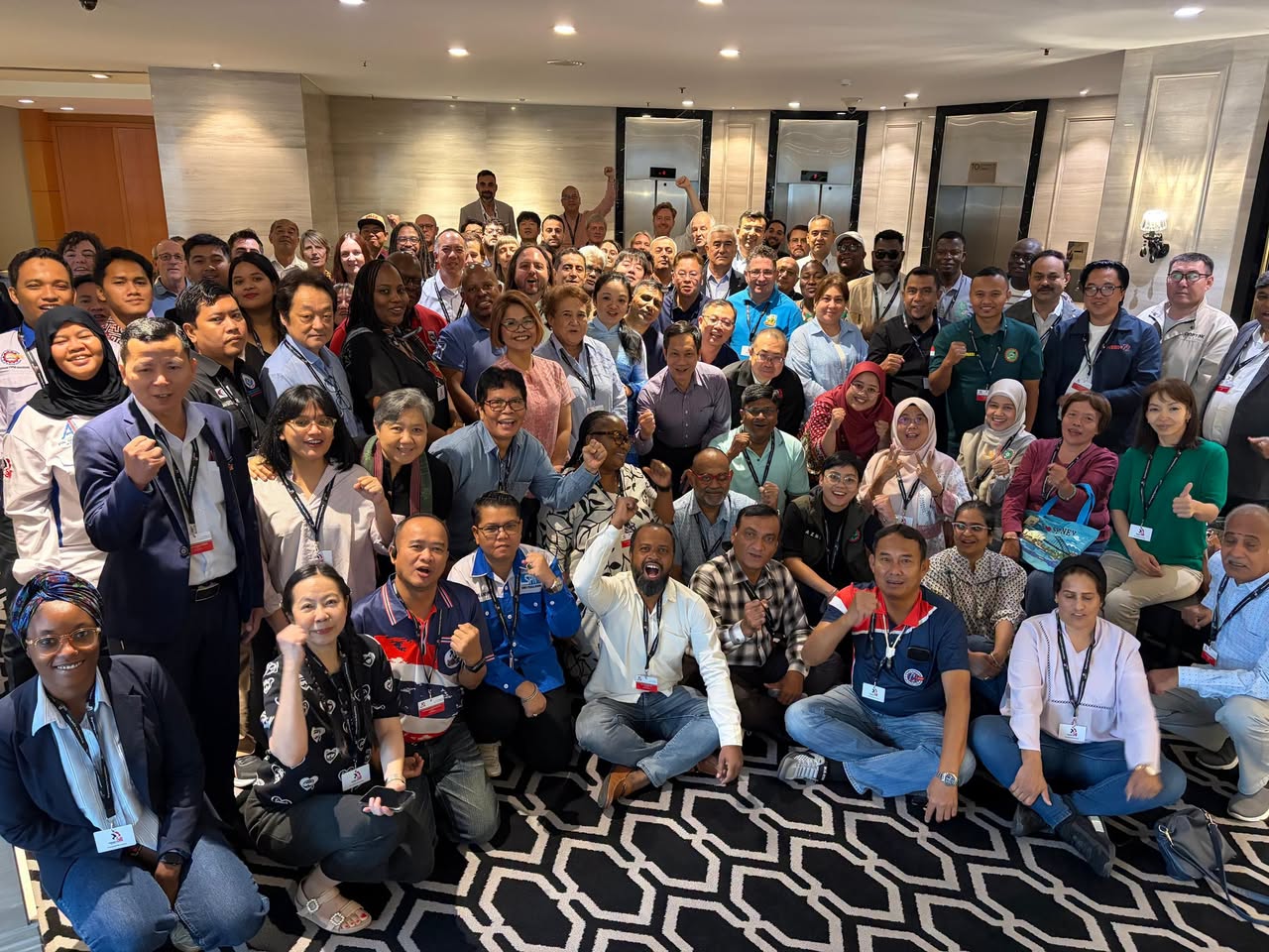 Large group of trade union delegates posing together in a hotel lobby, many raising their fists.