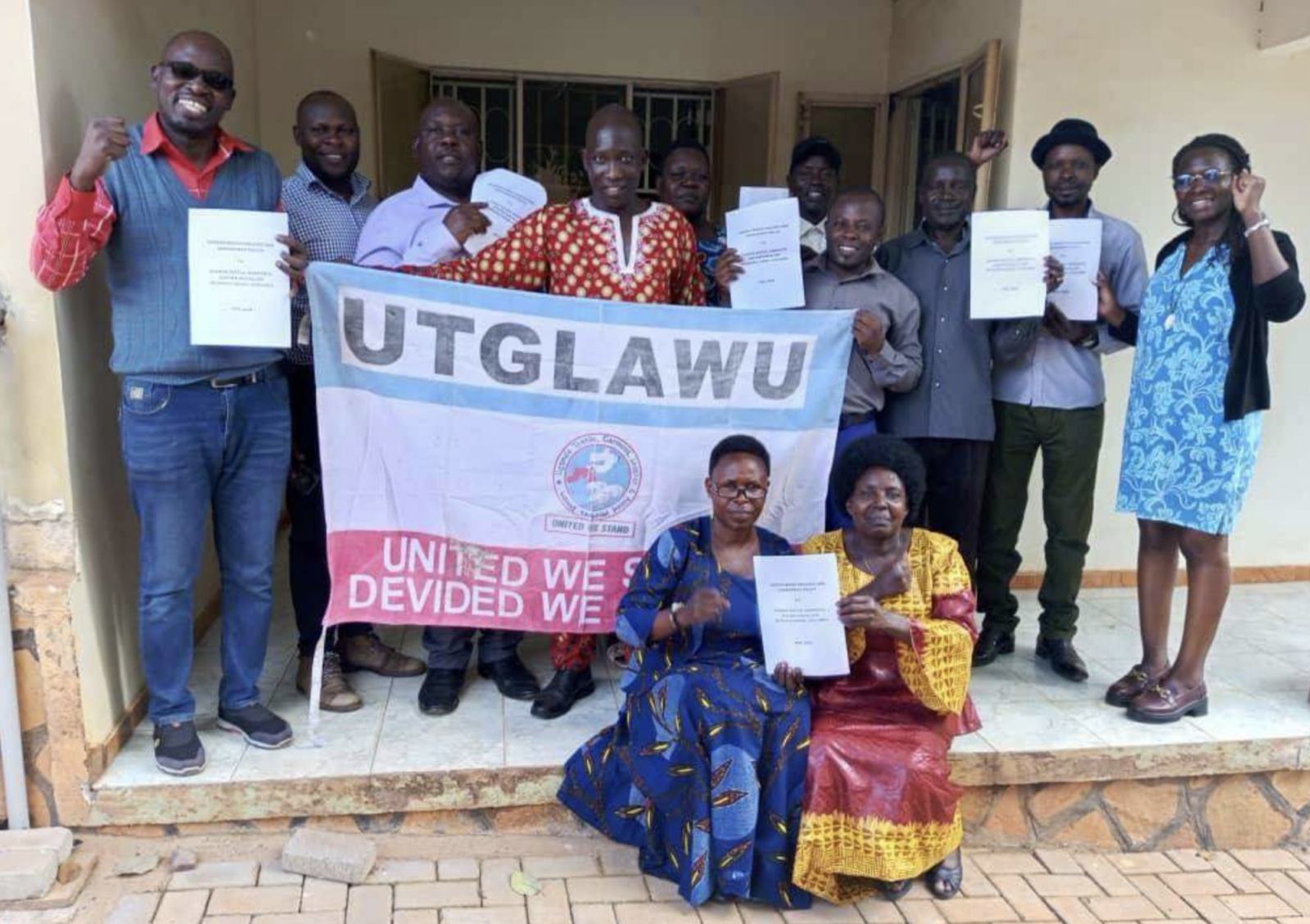 Members of the Uganda Textile, Garment, Leather and Allied Workers’ Union stand behind a union banner, holding up documents and raising their fists.