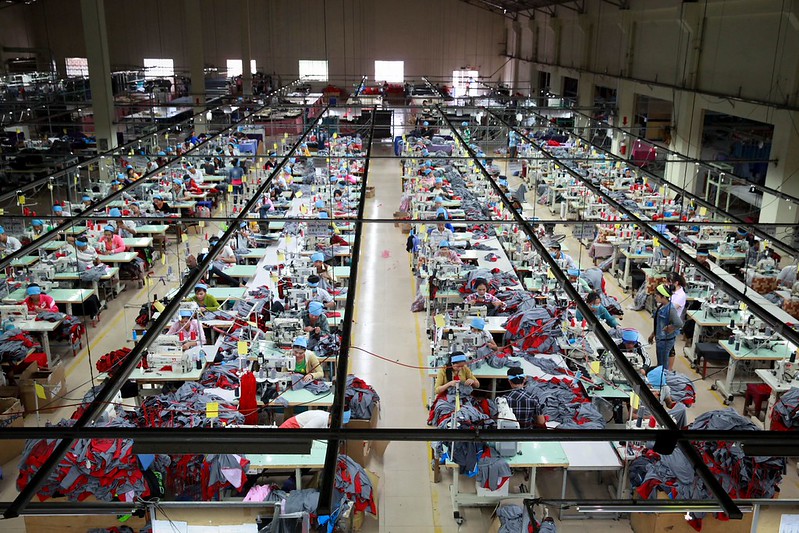 High-angle view of a large garment factory floor in Cambodia, with long rows of sewing machine stations where workers, mostly women in blue caps, sew piles of grey and red fabric.