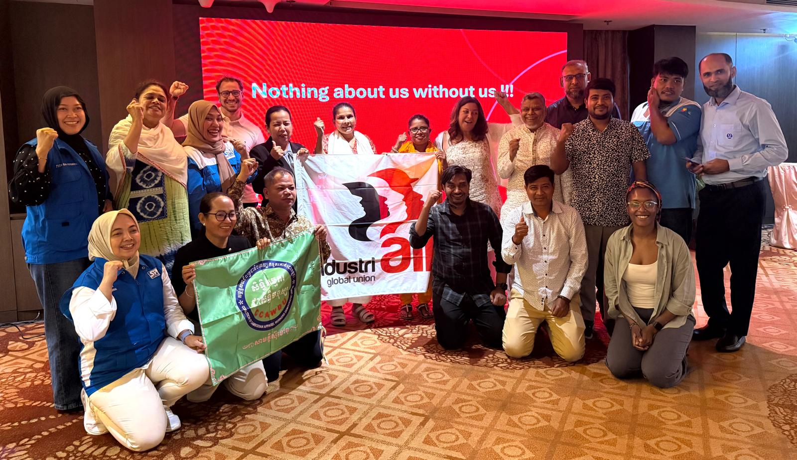 Union representatives from Bangladesh, Cambodia and Indonesia pose with raised fists, holding an IndustriALL Global Union flag and a C.CAWDU flag, in front of a red screen reading “Nothing about us without us!!!”