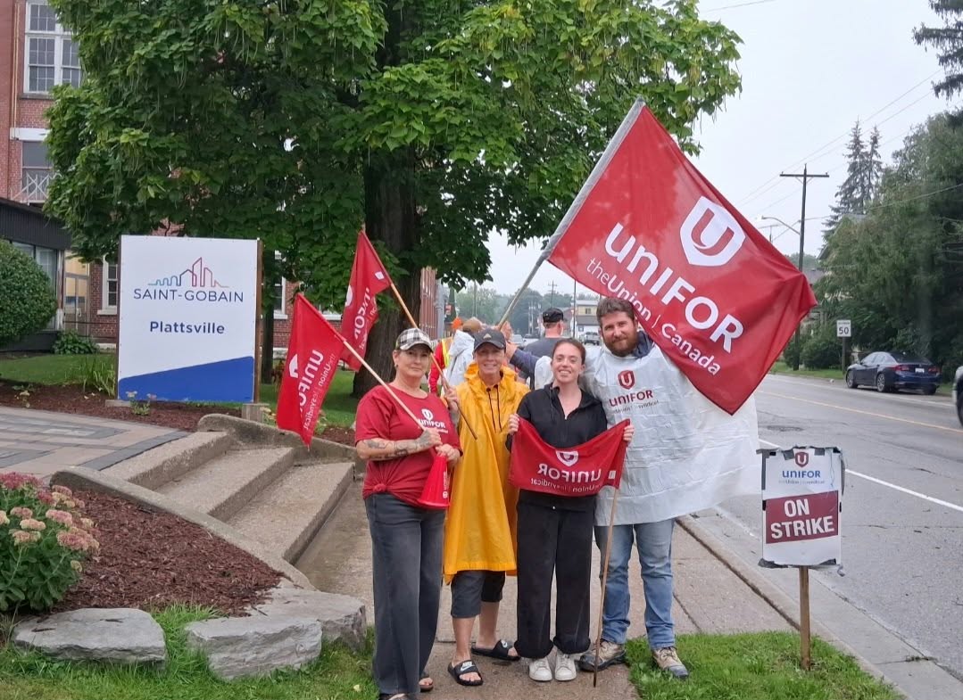 Cuatro miembros de Unifor con ropa de lluvia de pie sobre una acera mojada, sosteniendo banderas rojas de Unifor junto a un cartel de Saint-Gobain Plattsville y un cartel de piquete «On strike»