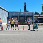 Cinco miembros de Unifor en fila sosteniendo carteles «On strike» frente a un almacén de metal azul con tres muelles de carga, conos de tráfico naranjas y una silla plegable vacía a su lado bajo un cielo azul despejado