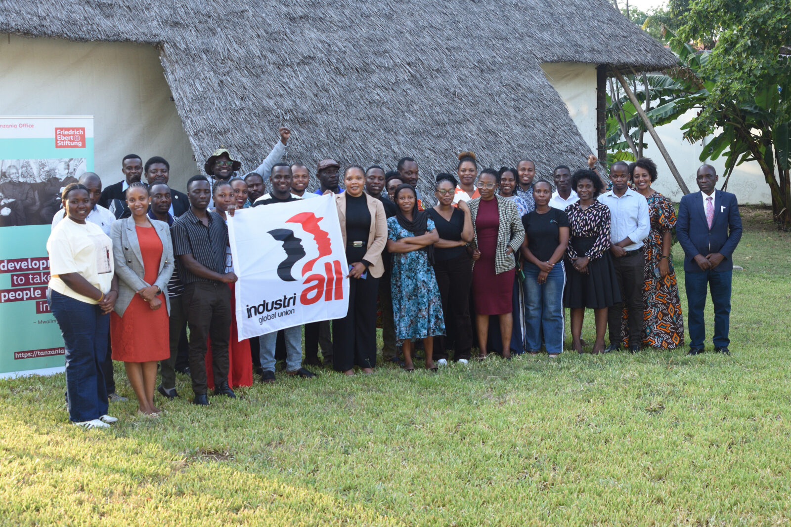 Group of young trade unionists holding an IndustriALL Global Union flag on a lawn, beside a Friedrich Ebert Stiftung Tanzania banner and a thatched building.