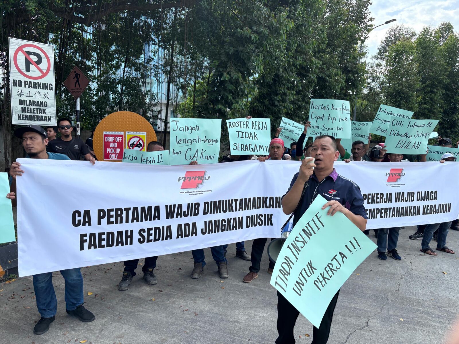 Workers stand behind a long white PPPMEU banner at a factory entrance, holding handwritten signs in Malay.