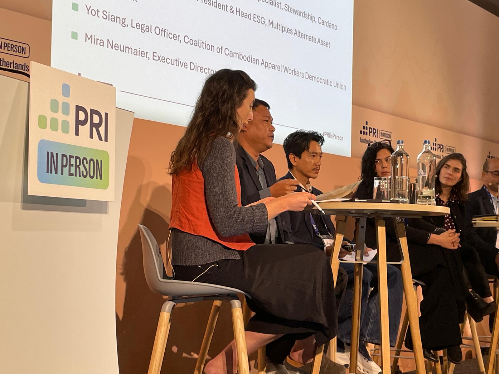 Six panellists seated on stage at PRI in Person in Amsterdam, with a PRI In Person sign on the left and a presentation screen behind them.